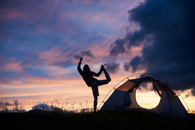 Sylwetka kobiety robi joga na szczycie góry na zachód słońca w pobliżu namiotu