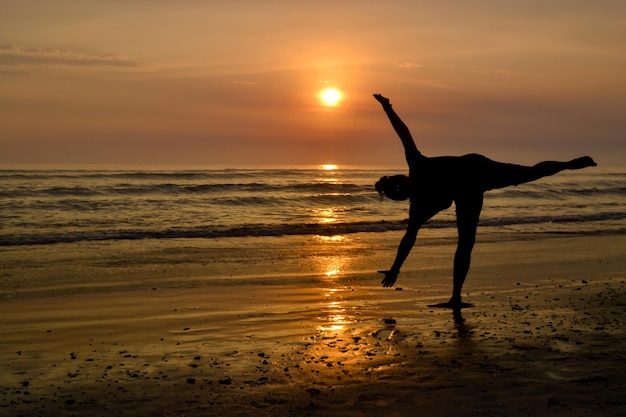 Sylwetka kobiety z otwartymi ramionami i na jednej nodze na brzegu plaży o zachodzie słońca