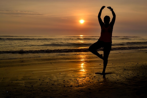 Sylwetka kobiety z otwartymi ramionami i na jednej nodze na brzegu plaży o zachodzie słońca