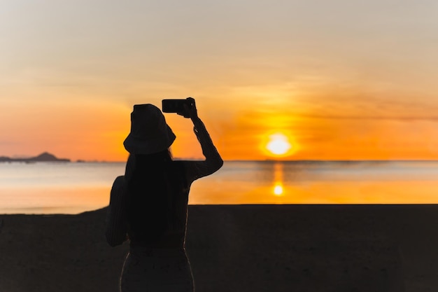 Sylwetka kobiety z telefonem komórkowym robiąca zdjęcie zachodu słońca na plaży