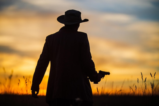 Sylwetka kowboja w kapeluszu z rewolwerem na dramatycznym niebie o zachodzie słońca Koncepcja zachodnia Życie na dzikim zachodzie