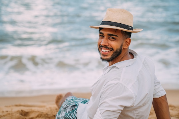 Sylwetka Młodego Człowieka Na Plaży. Latynoamerykański Mężczyzna Siedzi Na Piasku Plaży, W Kapeluszu, Patrząc W Kamerę