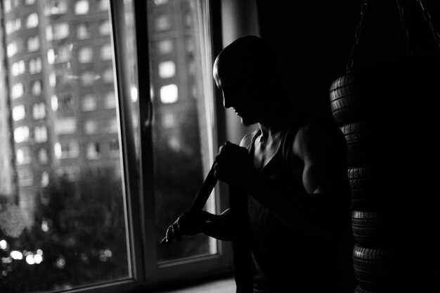 Sylwetka nie do poznania muskularny bokser owijający taśmę bokserską wokół ramion przed walką w centrum fitness z ciemnym wnętrzem. Mocna łysa zapalniczka przygotowująca się do noszenia rękawiczek przy oknie.