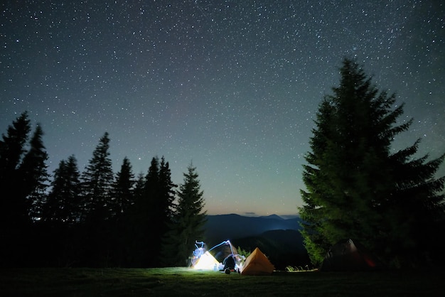 Sylwetka samotnego turysty odpoczywającego oprócz palenia ogniska w pobliżu oświetlonych namiotów turystycznych na kempingu w ciemnych górach pod nocnym niebem z gwiazdami. Aktywny styl życia i koncepcja życia na świeżym powietrzu.