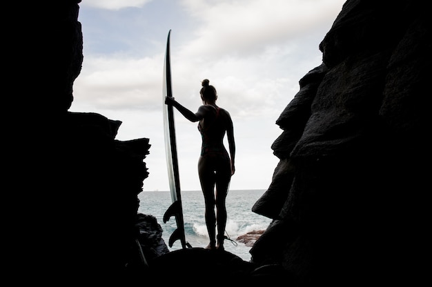 Sylwetka sportowy dziewczyna w strój kąpielowy stojący z surfowania wewnątrz skały na plaży Ocean Atlantycki patrząc w dół.