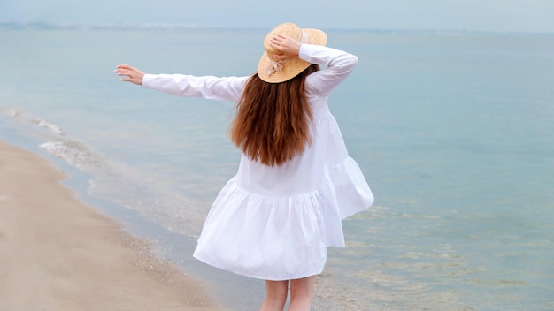 Sylwetka szczęśliwy cieszący się kobieta z tyłu z cienkimi nogami w białej sukni plażowej po odpoczynku o zachodzie słońca na plaży nad morzem Nie do poznania szczęśliwa kobieta w słomkowym kapeluszu w ręku na brzegu morza Długi baner internetowy