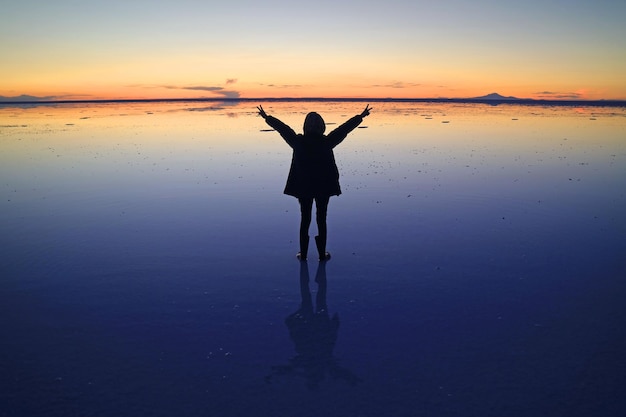 Zdjęcie sylwetka travellera wznosząca ramiona na lustrzanym efekcie uyuni salt flats o zachodzie słońca w boliwii