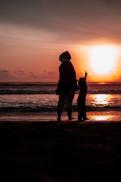 Sylwetki matki i córki trzymające się za ręce i wskazujące na zachód słońca na plaży