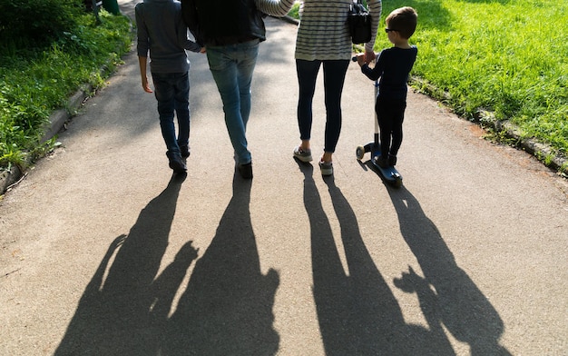 Sylwetki młodych ludzi w podświetleniu. Młody rodzinny tata, mama i dwoje dzieci spacerują ścieżką w parku w promieniach zachodzącego słońca