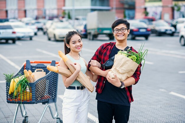 Sympatyczny azjatykci para trzyma papierowe eco torby z organicznie zdrowym jedzeniem w rękach podczas gdy stojący blisko sklepu centrum handlowego.