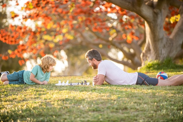 Szach-mat spędzający razem czas na zajęciach strategicznych i taktycznych tata i chłopiec grają w grę logiczną ojciec i syn grają w szachy na trawie w parku dzień ojca szczęśliwa rodzina rodzicielstwo i dzieciństwo