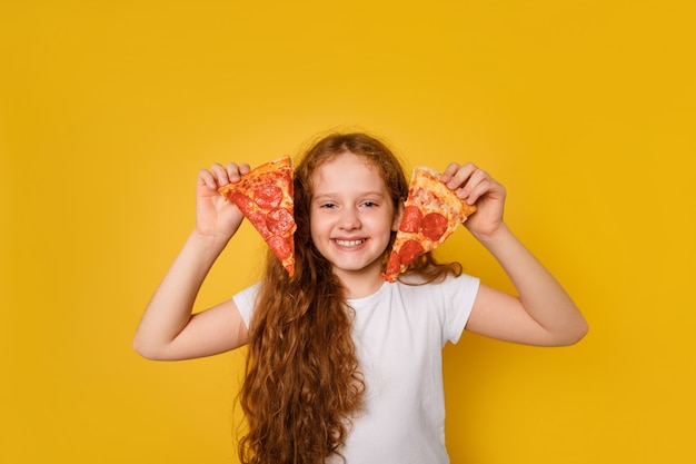 Szalona kędzierzawa dziewczyna trzyma dwa plasterki pizzy w pobliżu oczu i wystaje jej język.