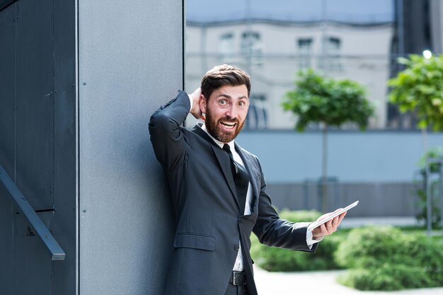 Szalony Niezadowolony Biznesmen Denerwuje Się Za Pomocą Inteligentnego Telefonu Cyfrowego Tabletu. Zły Człowiek Biznesu Ma Problem Z Mobilną Wiadomością Spamową. Mężczyzna Sfrustrowany Słabą Baterią Lub Złym Sygnałem W Biurze Na Zewnątrz