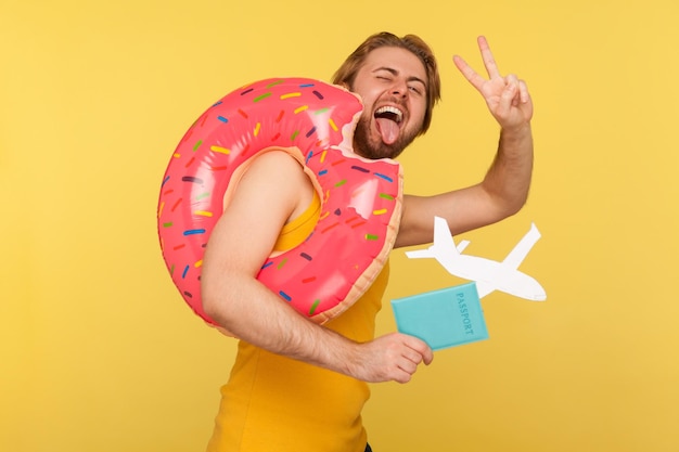 Szalony optymistyczny turysta w podkoszulku stojący z różowym gumowym pierścieniem pączka trzymającego dokument paszportowy i makieta samolotu pokazująca znak zwycięstwa v gest sukcesu wygrana studio strzał na białym tle