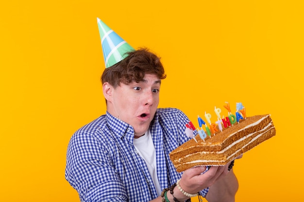 Zdjęcie szalony wesoły młody człowiek w papierowym gratulacyjnym kapeluszu trzyma ciasta wszystkiego najlepszego z okazji urodzin stojąc na żółtej ścianie. koncepcja jubileuszowe gratulacje.