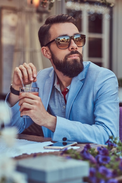 Szczegół portret modnego brodatego mężczyzny ze stylową fryzurą, trzyma kieliszek cappuccino, siedząc w kawiarni na świeżym powietrzu.