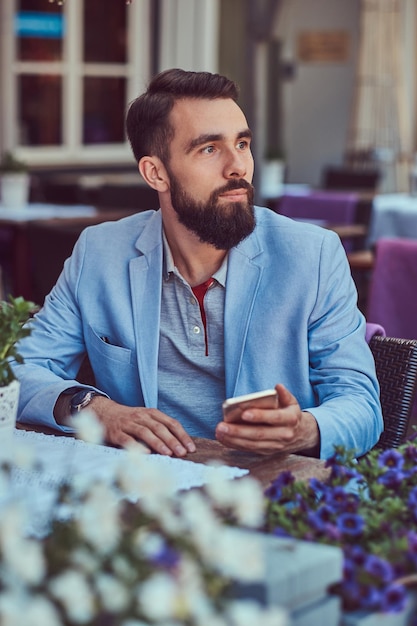 Szczegół portret modnego brodatego mężczyzny ze stylową fryzurą, trzyma smartfon, siedząc w kawiarni na świeżym powietrzu.