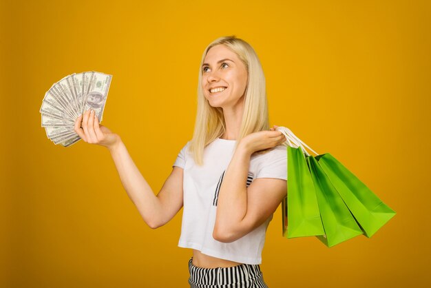 Zdjęcie szczegół portret szczęśliwa młoda piękna blondynka trzyma pieniądze i zielone torby na zakupy, na żółto