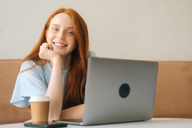 Szczegół portret wesoły młoda piękna kobieta siedzi przy biurku z laptopem i filiżanką z gorącą kawą w przytulnej kawiarni, patrząc na kamery. Ładna ruda kaukaska pani zdalna praca lub nauka.