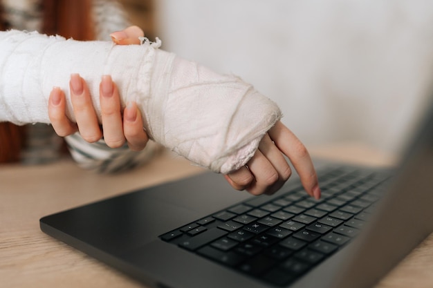 Szczegółowo przycięte ujęcie nierozpoznawalnej rannej freelancerki ze złamaną prawą ręką owiniętą w biały bandaż gipsowy pracujący wpisując na laptopie
