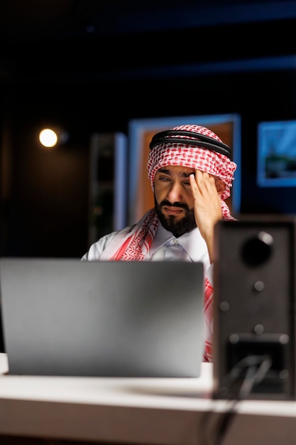 Szczegółowy widok zmęczonego mężczyzny w arabskim stroju siedzącego przy biurku i korzystającego z laptopa do prowadzenia badań w Internecie. Muzułmanin z minikomputerem na stole, wyglądający na zmęczonego i cierpiącego na ból głowy.