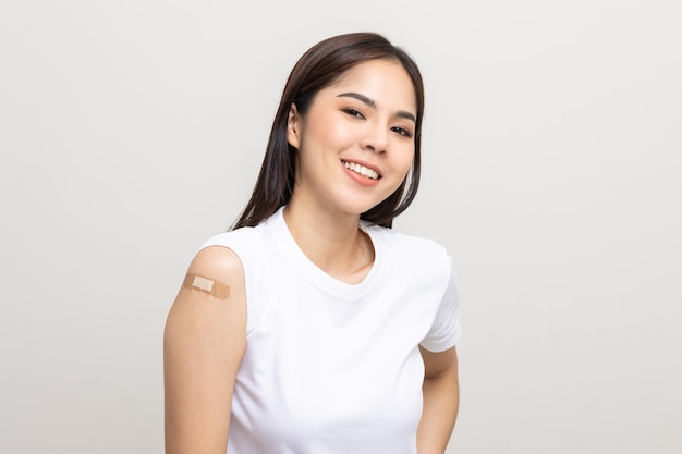 Szczepionka. Młoda piękna azjatycka kobieta dostaje szczepionkę chroniącą przed koronawirusem. Uśmiechnięta szczęśliwa kobieta pokazując ramię z bandażem po otrzymaniu szczepienia. Na na białym tle.