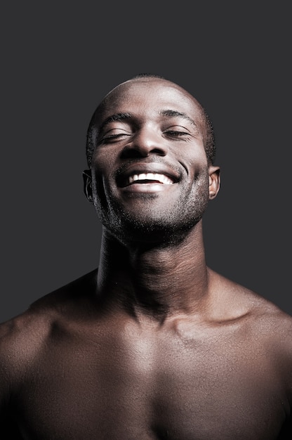 Szczere szczęście. Portret młodego bez koszuli afrykańskiego mężczyzny, który ma zamknięte oczy i uśmiecha się, stojąc na szarym tle