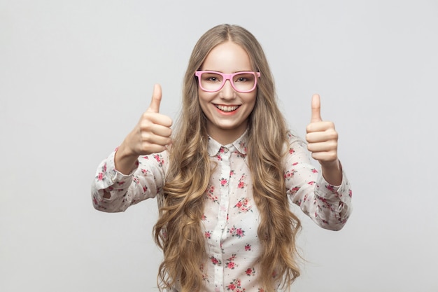 Szczęście Blondynka W Okularach Przeciwsłonecznych, Pokazując Kciuk Do Góry, Patrząc Na Kamery I Uśmiechając Się. Strzał Studio. Na Białym Tle Na Szarym Tle