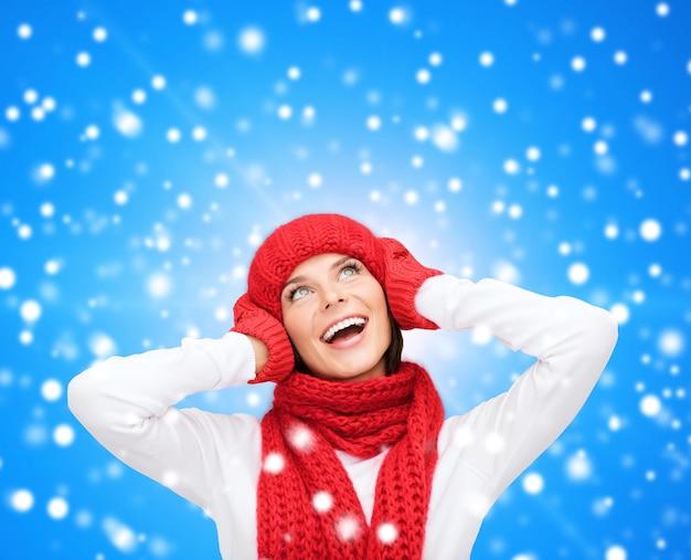 szczęście, ferie zimowe, święta i koncepcja ludzi - uśmiechnięta młoda kobieta w czerwonym kapeluszu, szaliku i rękawiczkach na niebieskim śnieżnym tle