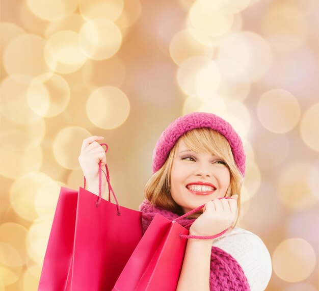 szczęście, ferie zimowe, święta i koncepcja ludzi - uśmiechnięta młoda kobieta w kapeluszu i szaliku z różowymi torbami na zakupy na beżowym tle światła