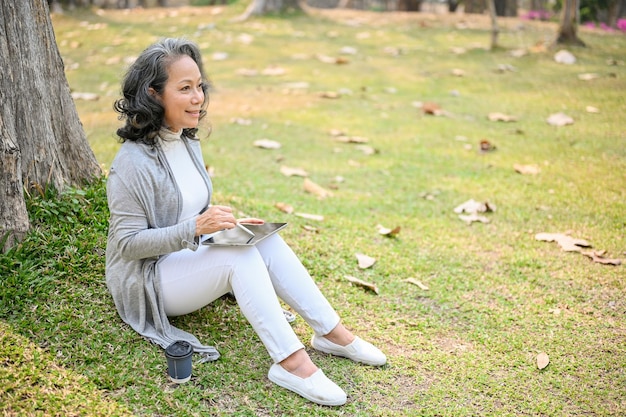 Szczęśliwa 60-letnia Azjatka używająca tabletu podczas relaksu i siedzenia na trawie pod