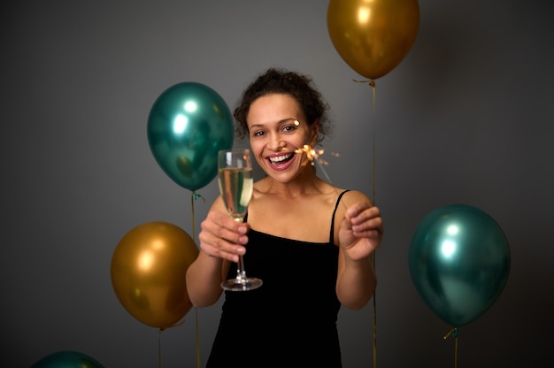 Szczęśliwa African American kobieta trzyma flet szampana i ognie, uśmiecha się z pięknym uśmiechem toothy poes na tle szarej ściany z napompowanymi złotymi i zielonymi balonami. Skopiuj miejsce na reklamę
