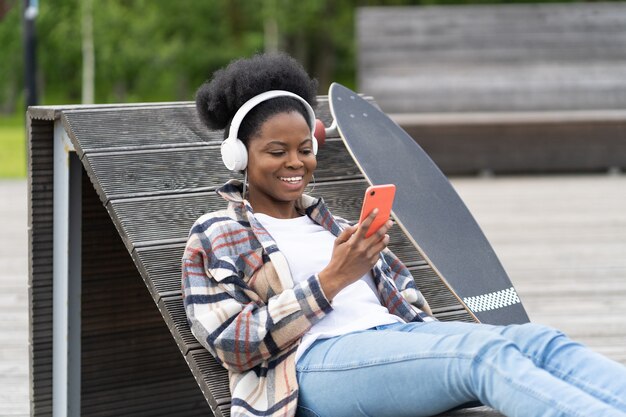 Szczęśliwa afroamerykanka z czatem na deskorolce na smartfonie siedzi w skate parku, wysyłając SMS-y na zewnątrz