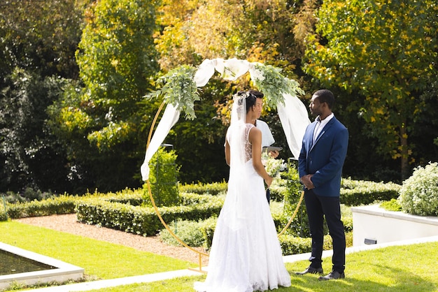 Szczęśliwa afrykańsko-amerykańska panna młoda i pan młody stoją pod łukiem ślubnym w słonecznym ogrodzie.