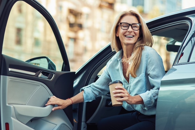 Szczęśliwa atrakcyjna kobieta lub biznes dama w okularach, trzymając filiżankę kawy i wysiadając z jej nowoczesny samochód, koncepcja transportu