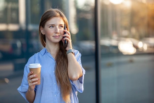 Szczęśliwa atrakcyjna uśmiechnięta kobieta, rozmawia przez telefon komórkowy, trzyma kawę na wynos