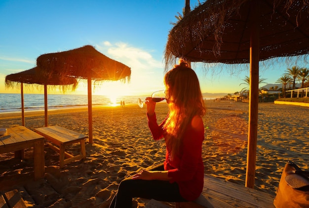 Szczęśliwa Azjatka pije czerwone wino i ogląda zachód słońca na plaży.