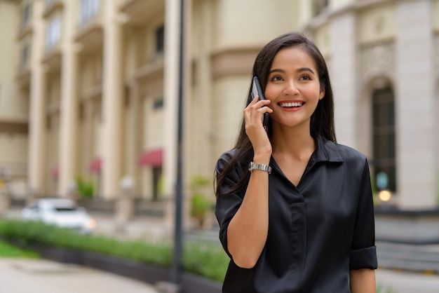 Szczęśliwa azjatycka bizneswoman na zewnątrz na ulicy miasta rozmawia przez telefon, uśmiechając się i spacerując