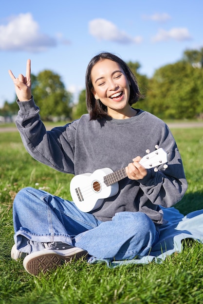 Szczęśliwa azjatycka dziewczyna grająca na ukulele w parku, pokazująca rock n roll heavy metal rogi znak i uśmiechnięty havi