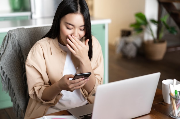 Szczęśliwa Azjatycka Dziewczyna Siedzi Z Laptopem W Domu, Patrząc Na Telefon Komórkowy I śmiejąc Się, Ma Miejsce...