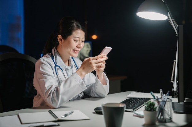 Szczęśliwa azjatycka japońska lekarka siedzi przy stole i przegląda stronę internetową na telefonie komórkowym późną nocą. piękna kobieta uśmiechający się personel pielęgniarki w ciemnym biurze kliniki przy użyciu telefonu komórkowego.