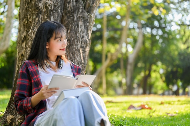 Szczęśliwa azjatycka kobieta czyta książkę i marzy o swoim sukcesie, siedząc pod drzewem