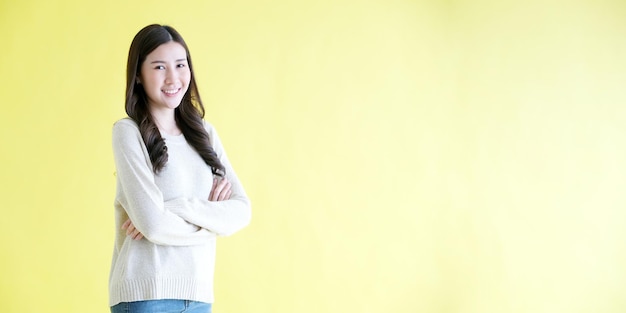 Szczęśliwa azjatycka kobieta krzyżuje ramię, uśmiechając się, stojąc nad odizolowanym żółtym tłem