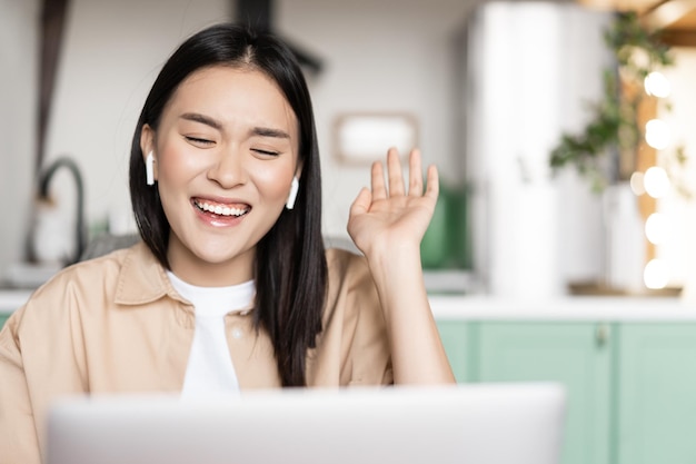 Szczęśliwa azjatycka kobieta śmiejąca się i uśmiechnięta rozmawiająca podczas wideorozmowy za pomocą laptopa machającego ręką i chichocząc ...
