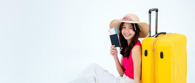 Szczęśliwa Azjatycka kobieta w różowej koszuli bez rękawów i kapeluszu na plaży, trzymając bilet lotniczy paszport i żółtą walizkę na białym tle transparent z miejsca kopiowania Młody podróżnik gotowy na letnią wycieczkę