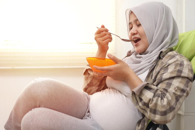 Zdjęcie szczęśliwa azjatycka muzułmańska mama w ciąży siedzi na krześle i je zdrowe jedzenie dla swojego dziecka.