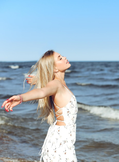 Szczęśliwa blond piękna kobieta na plaży oceanu stojąca w białej letniej sukience, z otwartymi ramionami