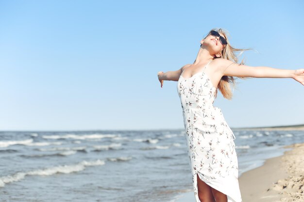 Szczęśliwa blondynka piękna kobieta na plaży oceanu stojąca w białej letniej sukience i okularach przeciwsłonecznych, otwarte ramiona.