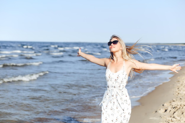 Szczęśliwa blondynka, piękna kobieta na plaży oceanu, stojąca w białej letniej sukience i okularach przeciwsłonecznych, z otwartymi ramionami.