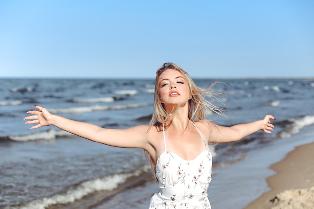 Szczęśliwa blondynka piękna kobieta na plaży oceanu stojąca w białej letniej sukience, podnosząc ręce.